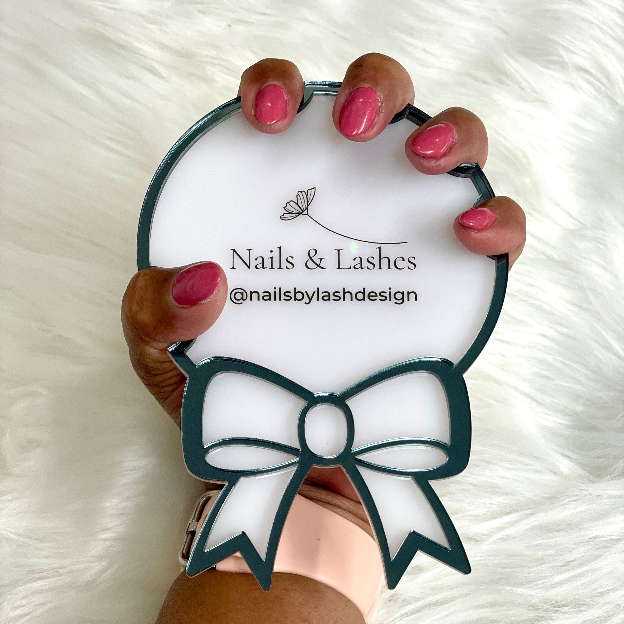 Hand holding a round mirror with 'Nails & Lashes' branding on a white fluffy background