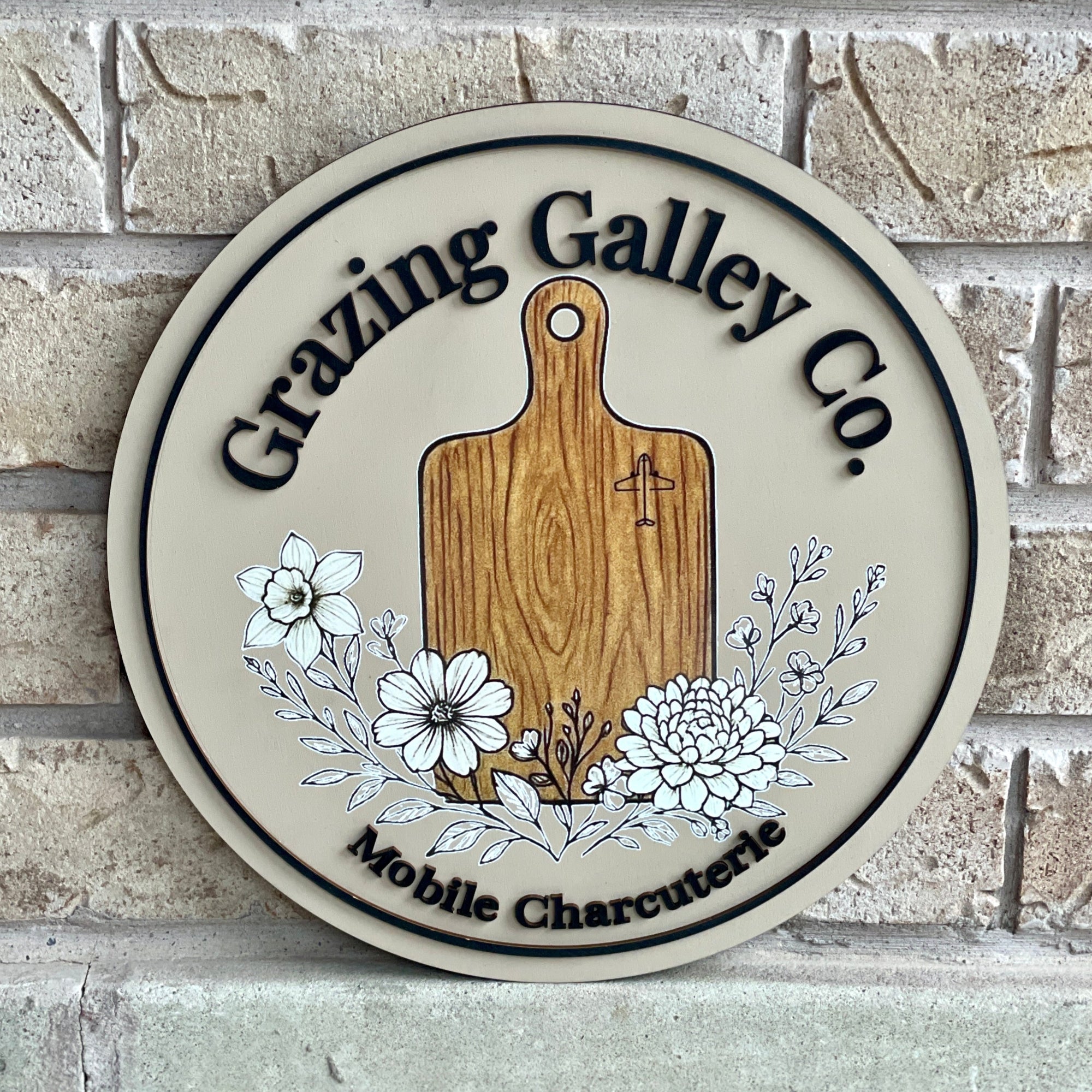 Round wooden sign with 'Grazing Galley Co.' branding on a brick wall background