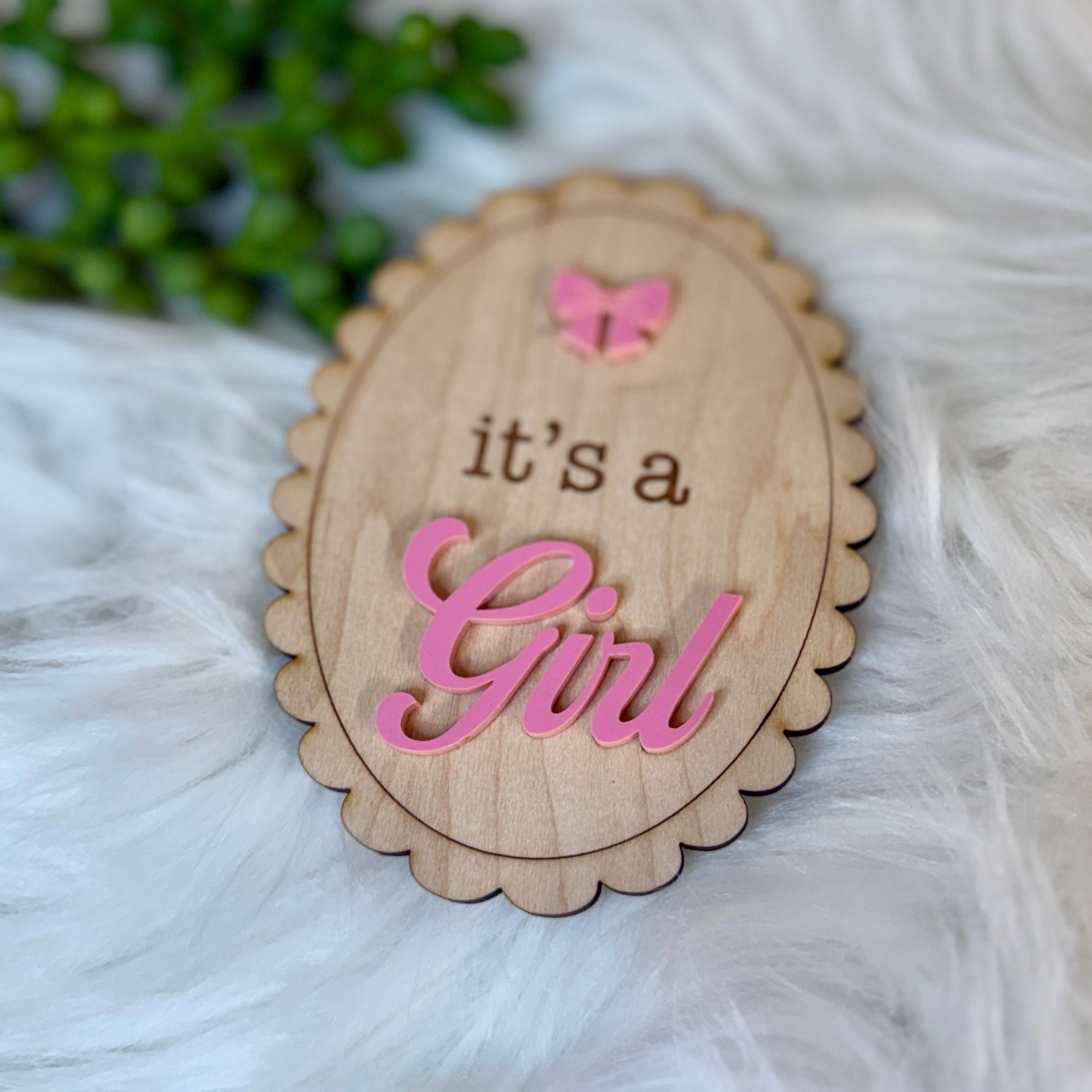 Wooden plaque with 'it's a Girl' text on a fluffy white surface with greenery.