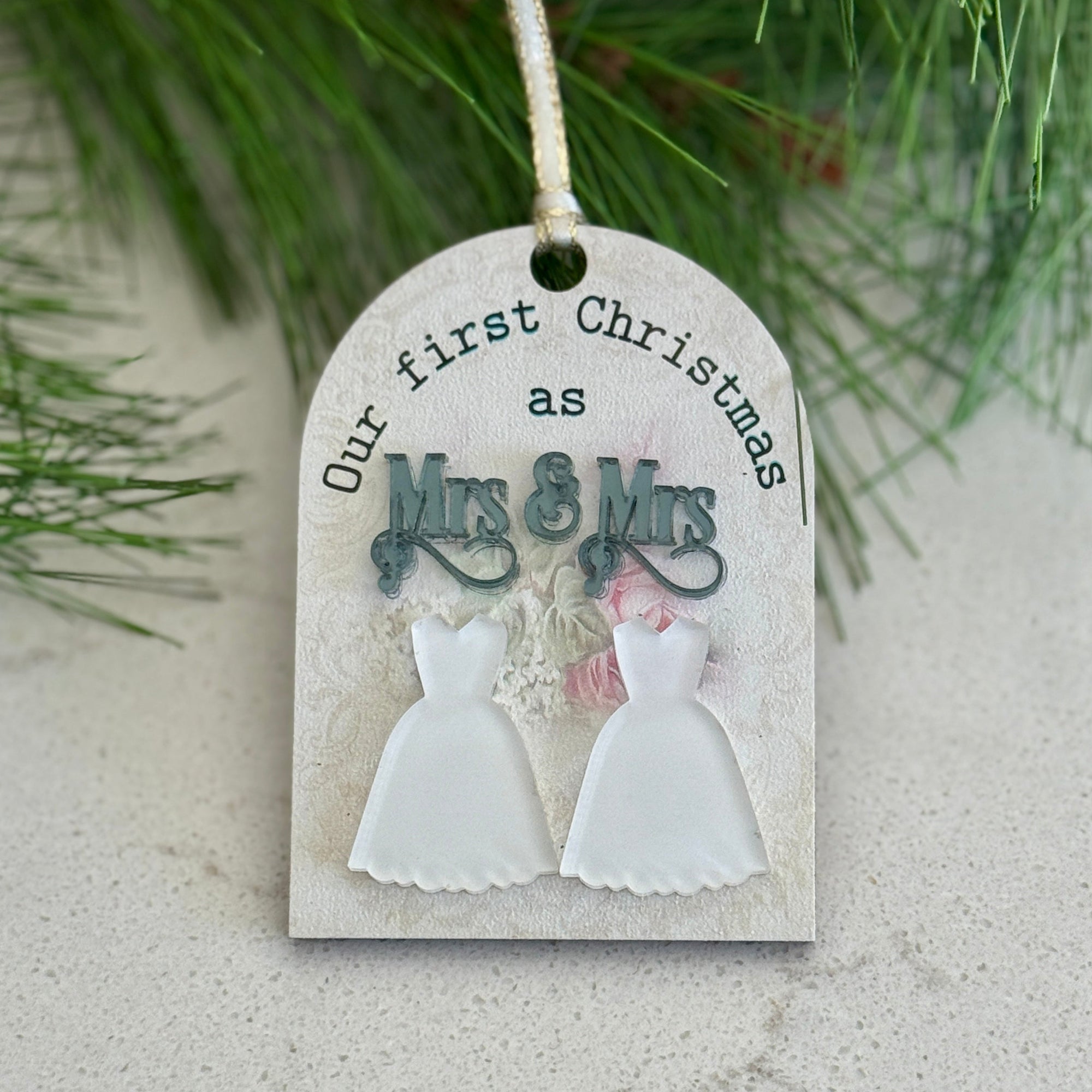 Decorative tag with 'Our first Christmas as Mrs & Mrs' text and wedding dress illustrations, hanging on a branch.