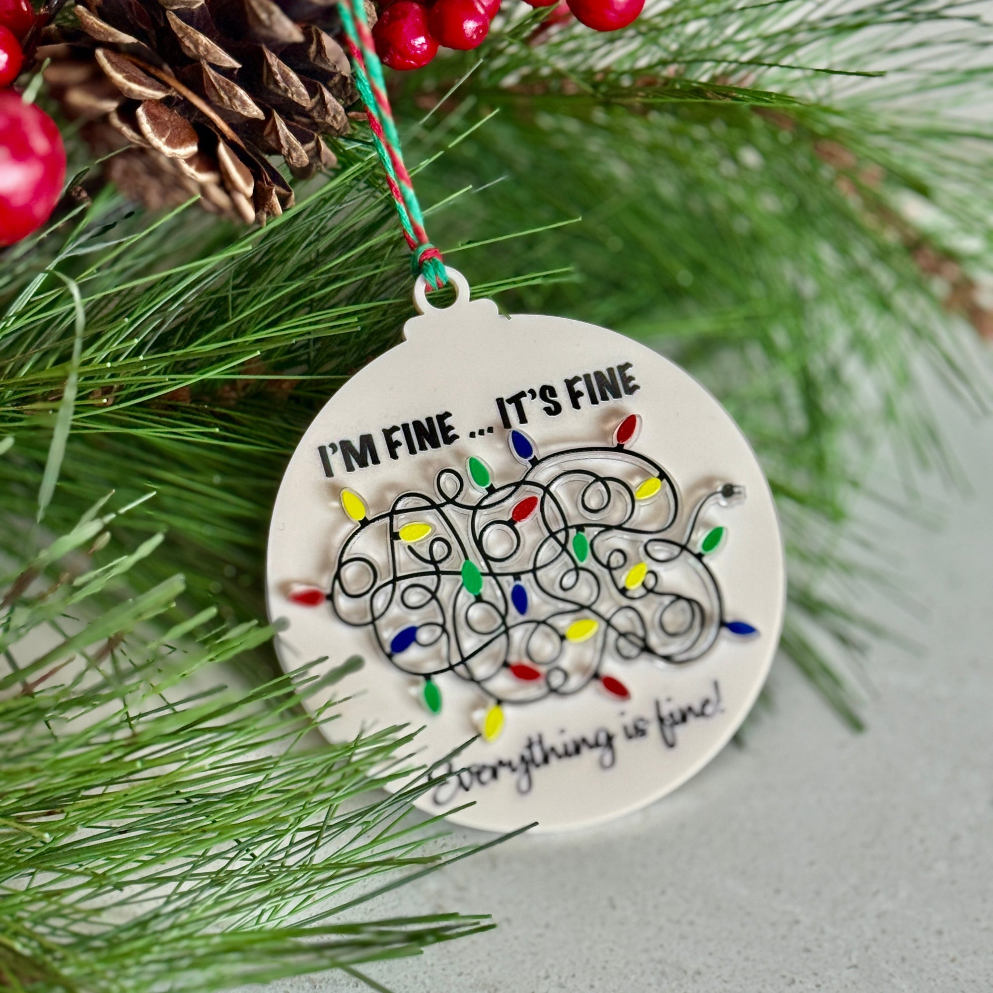 Christmas ornament with text 'I'm fine... it's fine, everything is fine' on a branch with pine needles and berries.