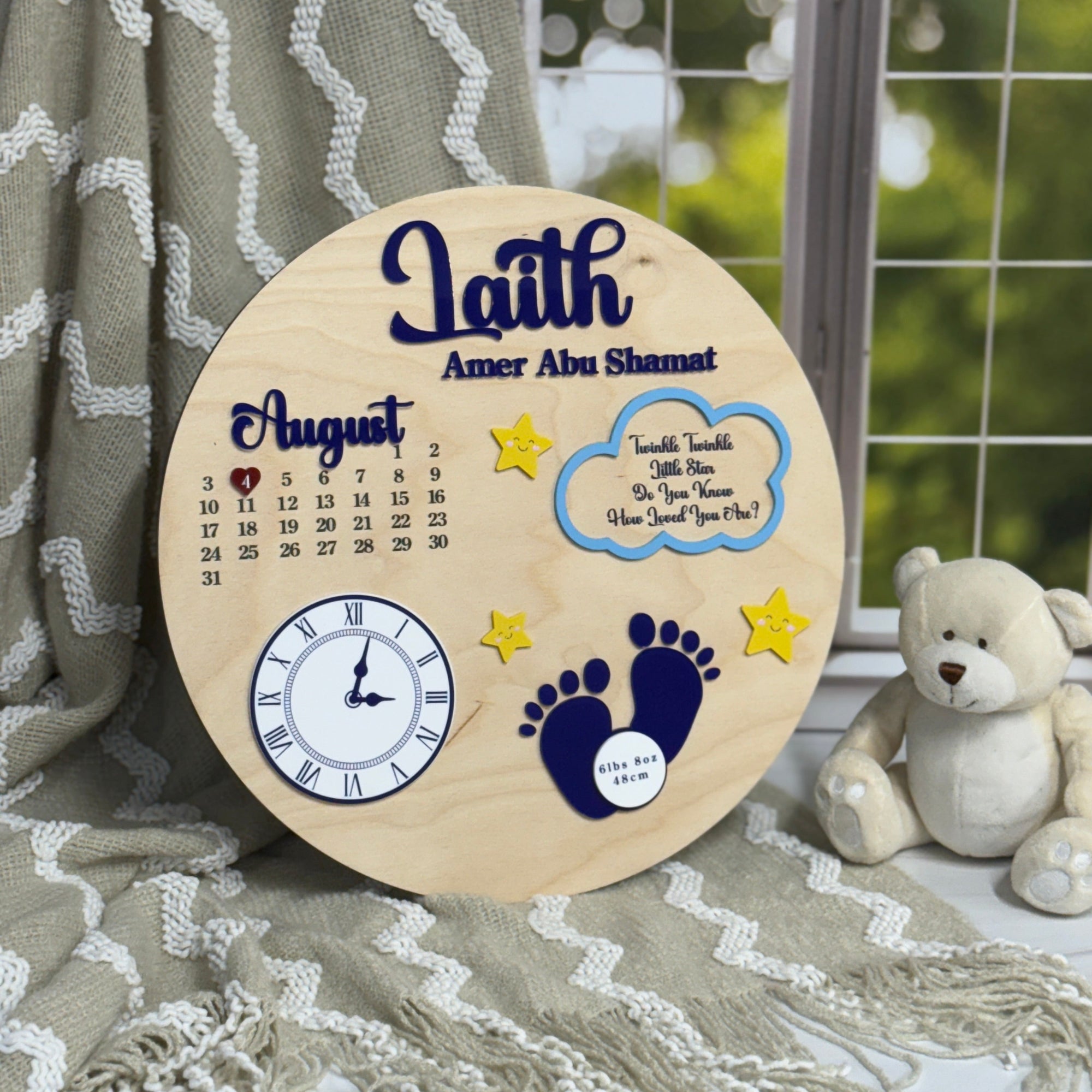 Wooden plaque with baby-themed designs and text, placed on a textured surface with a teddy bear in the background.