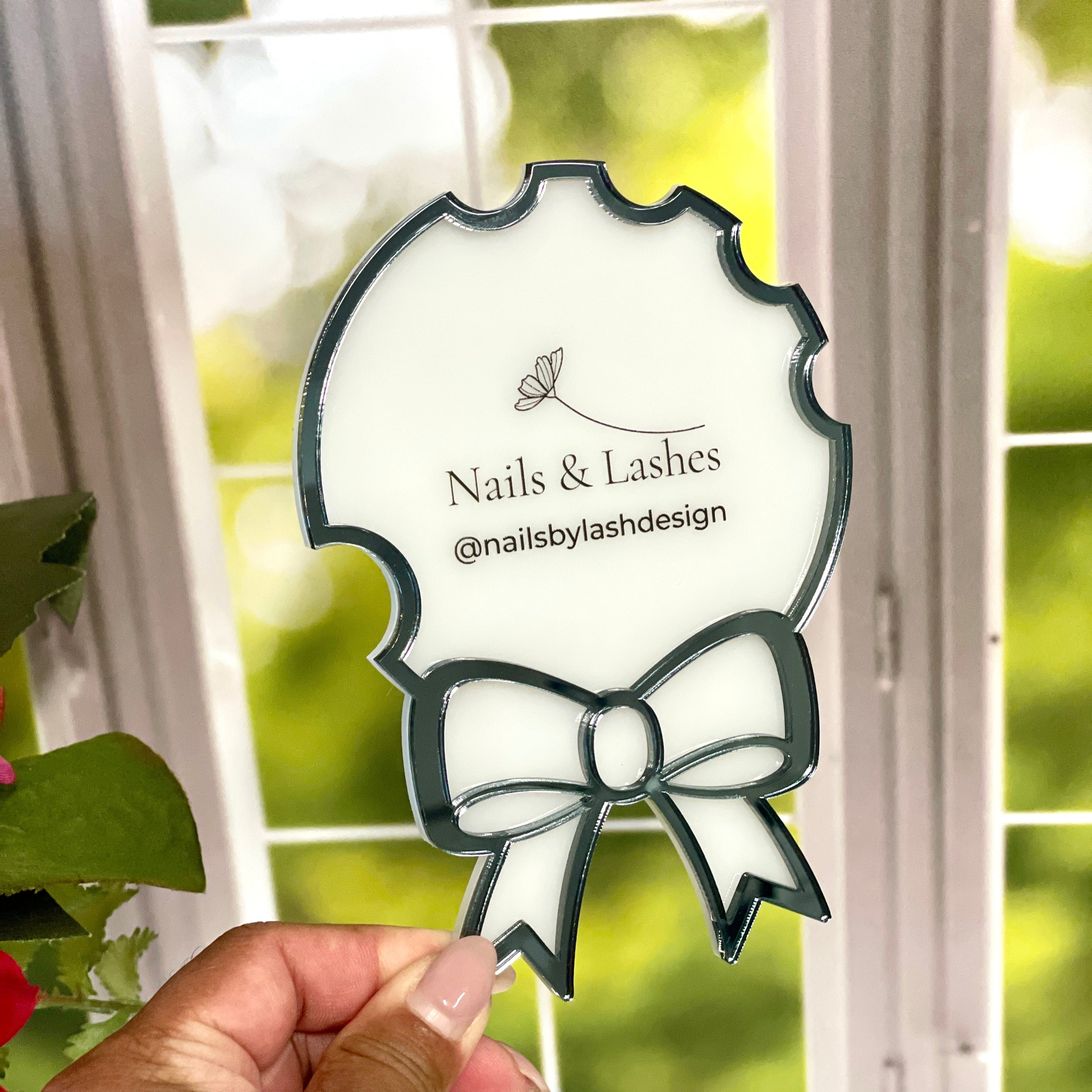 Hand holding a decorative mirror with 'Nails & Lashes' branding in front of a window with greenery.