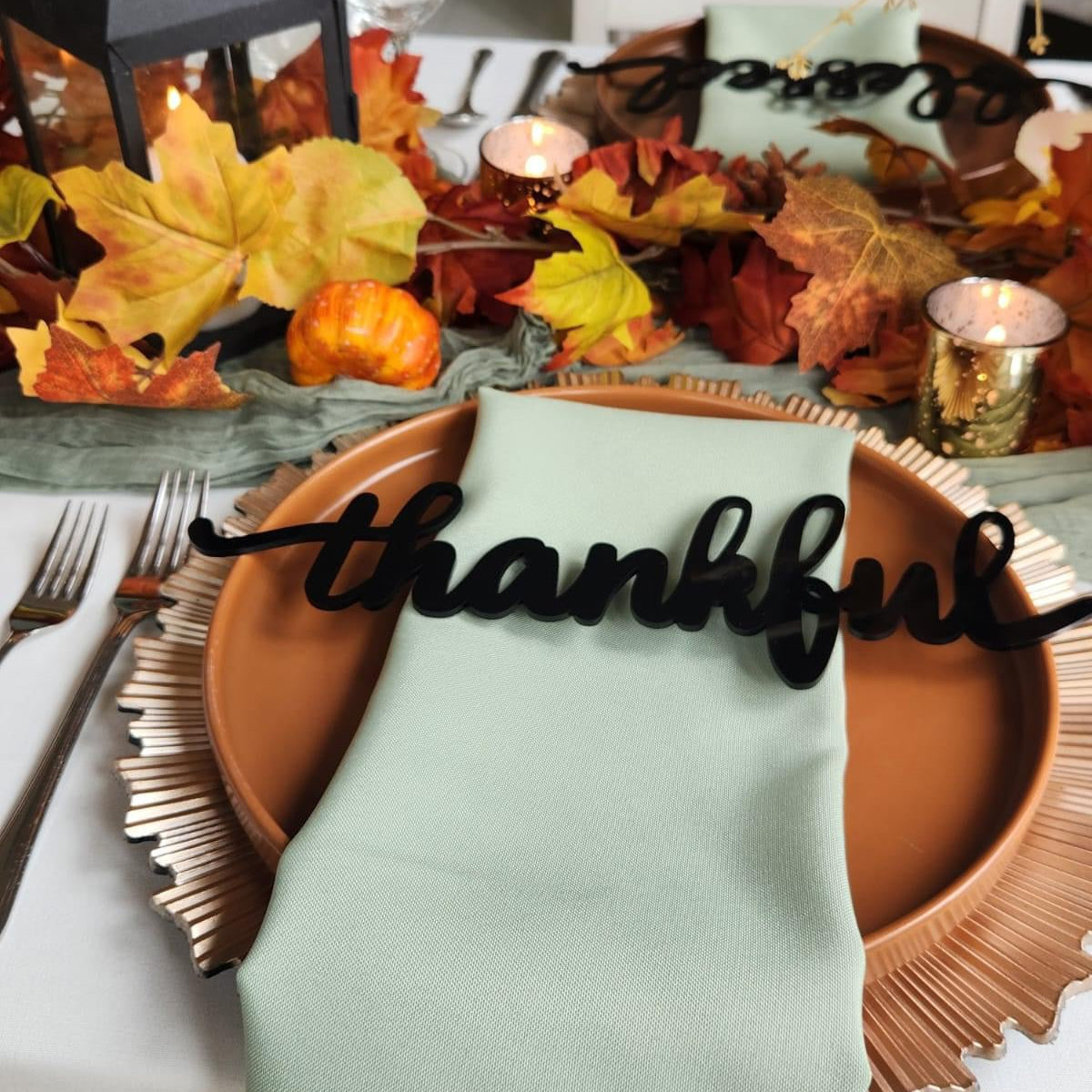 Place setting with a 'Thankful' themed place card on a round wooden plate, with fall-themed decorations in the background.