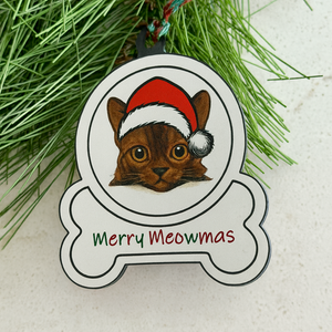 Burmese Cat ornament with Santa hat and 'Merry Meowmas' text on a white surface with greenery.