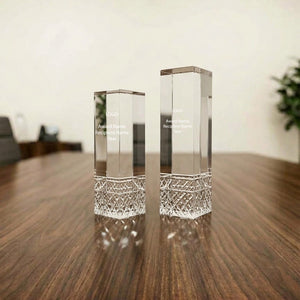 Two clear rectangular glass or crystal award plaques with engraved text and a faceted cut base, standing on a wooden table in a softly blurred indoor setting.