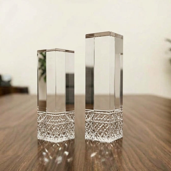 Two clear rectangular glass or acrylic display blocks with textured cut bases, standing on a dark wood table in a softly blurred indoor setting.