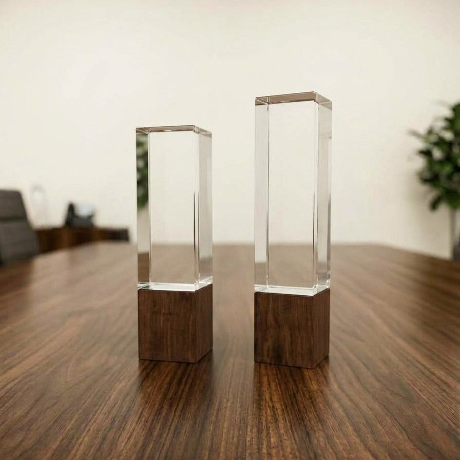 Two tall, minimalist rectangular pillar awards made of clear crystal mounted on solid dark walnut wood bases, displayed on a wooden table.