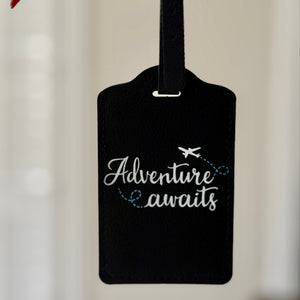 A black leather luggage tag with the phrase 'Adventure awaits' in white script and a small airplane graphic, held by a hand with red nail polish against a softly blurred indoor background.
