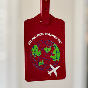 A red leather luggage tag with a gold buckle, held by a hand with red nail polish. The tag features a green and blue globe graphic with the text 'ALL YOU NEED IS A PASSPORT' and a small white airplane.