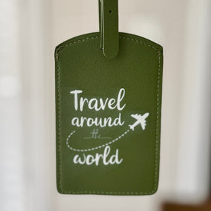 An olive green faux leather luggage tag with white text reading 'Travel around the world' and a small airplane graphic, held by a hand with red nail polish against a blurred indoor background.