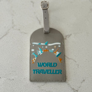 A gray leather luggage tag with a strap and buckle, featuring a colorful graphic of travel icons and the text 'WORLD TRAVELLER' in teal, placed on a light gray marble surface.