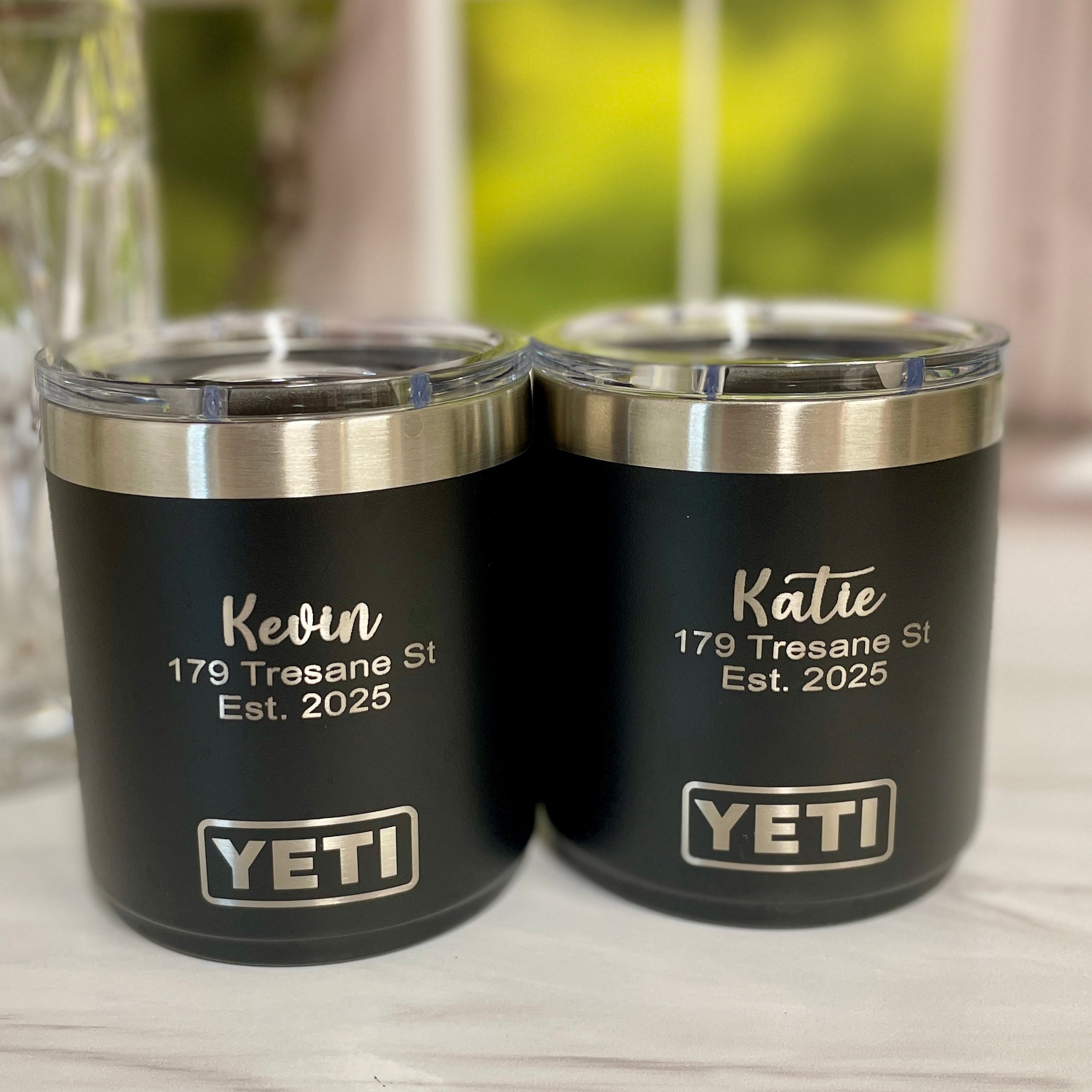 Two YETI tumblers with personalized names and addresses on a wooden surface.