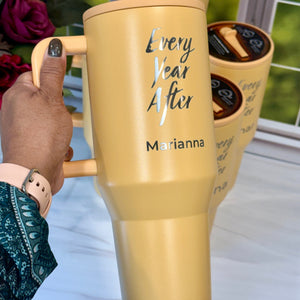 Yellow mug with 'Every Year After Marianna' text held by a hand, with another similar mug in the background.