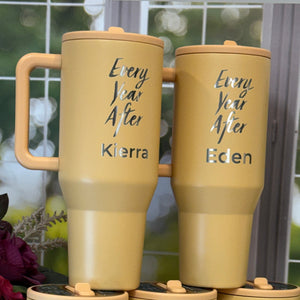 Gold tumblers with names engraved on them, placed against a blurred outdoor background.