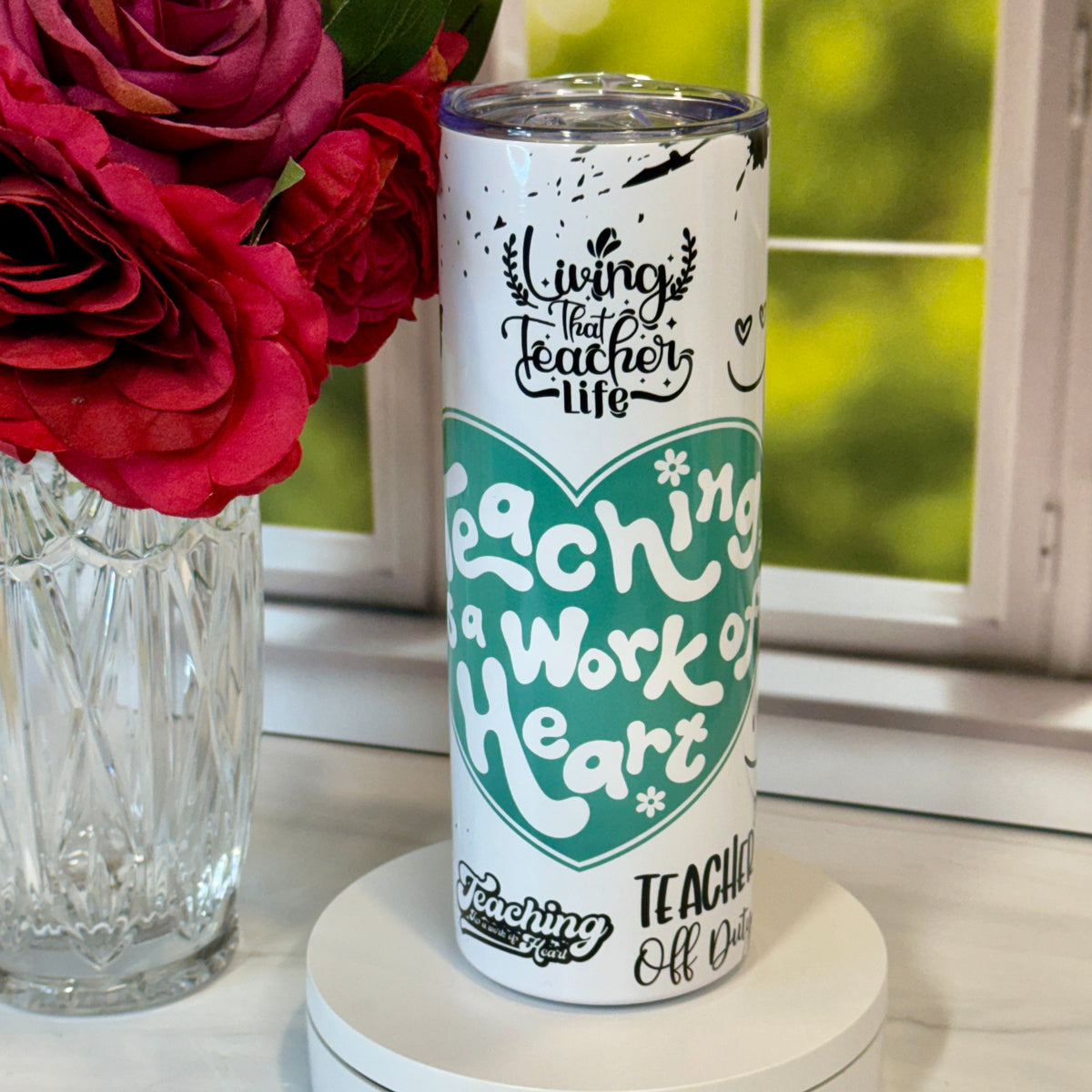 Tumbler with 'Teaching is a Work of Heart' design on a white surface with red flowers in the background.