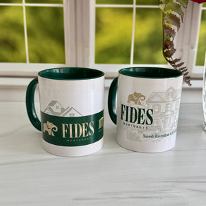 Two white ceramic mugs with dark green interiors and handles sit on a light marble countertop in front of a window. The left mug has a dark green band with gold FIDES Mortgage lettering and house illustrations, and the right mug has a pale gray house illustration with large FIDES Mortgage lettering.