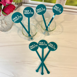 Five teal and white wooden cocktail picks with circular 'VAN J. HOUTTE' labels, shown with three in small clear glasses and two standing on a light wood tabletop near a window and flowers.