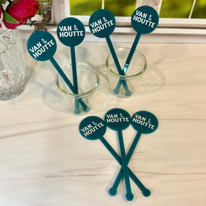 Six teal cocktail straws with round dark teal labels reading 'VAN 1/2 HOUTTE' in white text, arranged on a light tabletop near a window with flowers in the background.