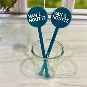 Two teal Van Houtte branded stirrers in a small clear glass cup, set on a light tabletop with a window and greenery blurred in the background.