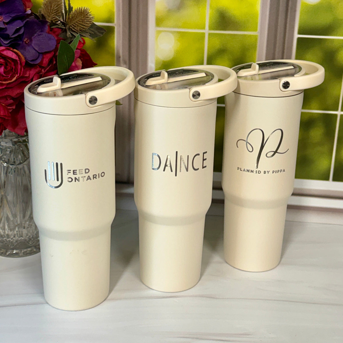 Three cream-colored insulated travel tumblers with lids are lined up on a light marble surface in front of a window with a blurred green garden background. The tumblers feature black printed text and logos: 'FEED ON TARIO' on the left, 'DANCE' in the center, and a cursive monogram with 'PLANNED BY HOP' on the right.