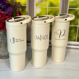 Three cream-colored insulated travel tumblers with lids are lined up on a light marble surface in front of a window with a blurred green garden background. The tumblers feature black printed text and logos: 'FEED ON TARIO' on the left, 'DANCE' in the center, and a cursive monogram with 'PLANNED BY HOP' on the right.