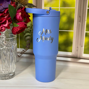 A light blue insulated travel tumbler with a matching lid sits on a white windowsill next to a glass vase of flowers. The tumbler is printed with the text 'Miss Mindy' in metallic gold script.