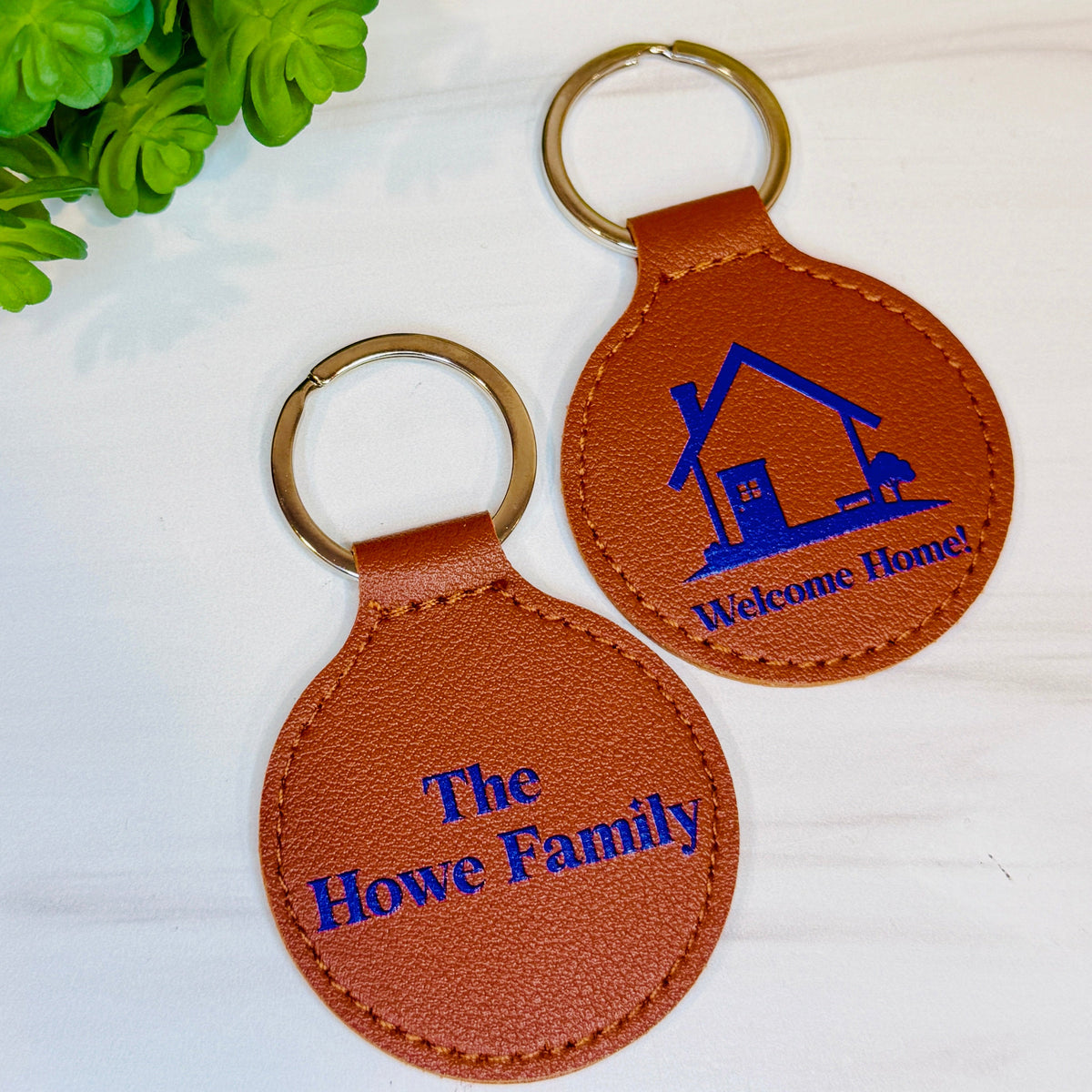 Two personalized brown leather keychains with brass key rings on a white surface, one featuring the text 'The Howe Family' and the other 'Welcome Home!' with a house graphic, next to a green plant.