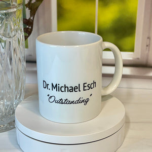 A white ceramic mug with black text reading 'Dr. Michael Esch *Outstanding*' sits on a white circular base on a tabletop.