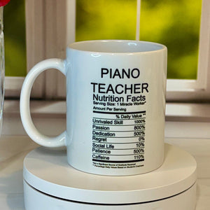 A white ceramic mug with a black printed nutrition facts label design and the words 'PIANO TEACHER' on the side, displayed on a white circular stand on a light wood surface next to a vase of red roses.