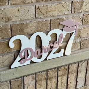 A decorative wooden sign spelling out '2027' with the name 'Darcel' in pink script across the center and a small pink graduation cap at the top, mounted on a tan brick wall.