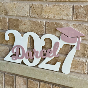White 2027 personalized wall sign with the name 'Darcel' in pink script and a pink graduation cap, mounted on a tan brick wall.