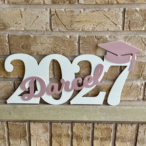 A decorative wooden sign spelling '2027 Darcel' with a pink graduation cap, mounted on a tan brick wall.