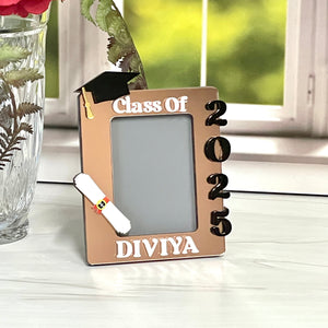 Graduation-themed photo frame with 'Class of 2025' and 'DIVIYA' on a white surface with a blurred window background.