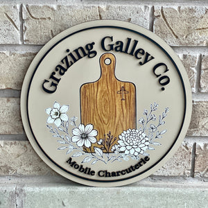 Round wooden sign with 'Grazing Galley Co.' branding on a brick wall background