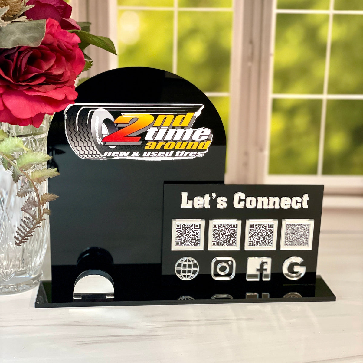 Black stand with QR codes and social media icons on a white surface with flowers in the background