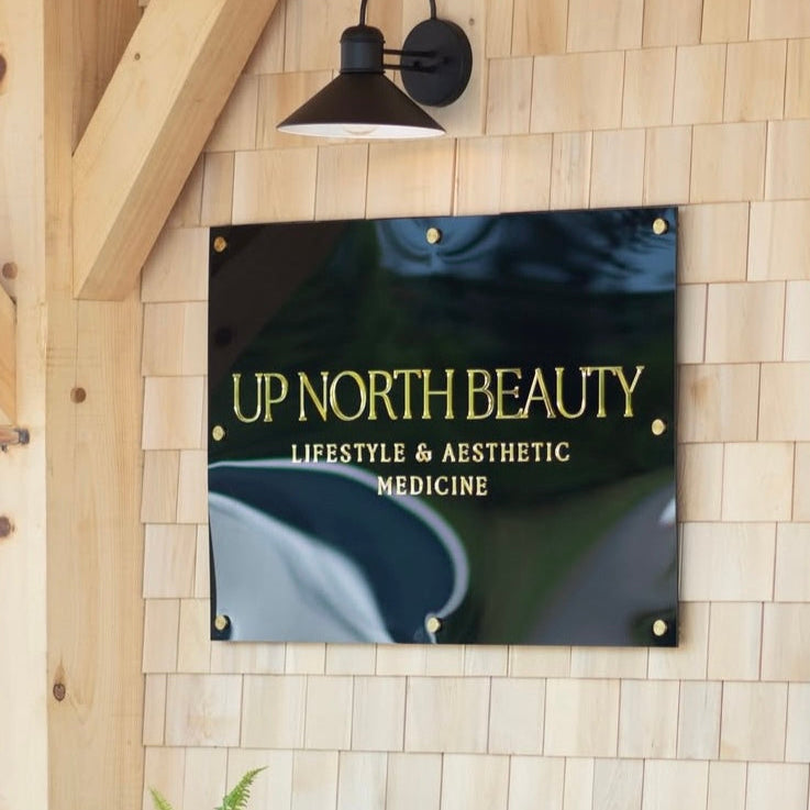 Glass sign with 'Up North Beauty' on a tiled wall with a potted plant below.