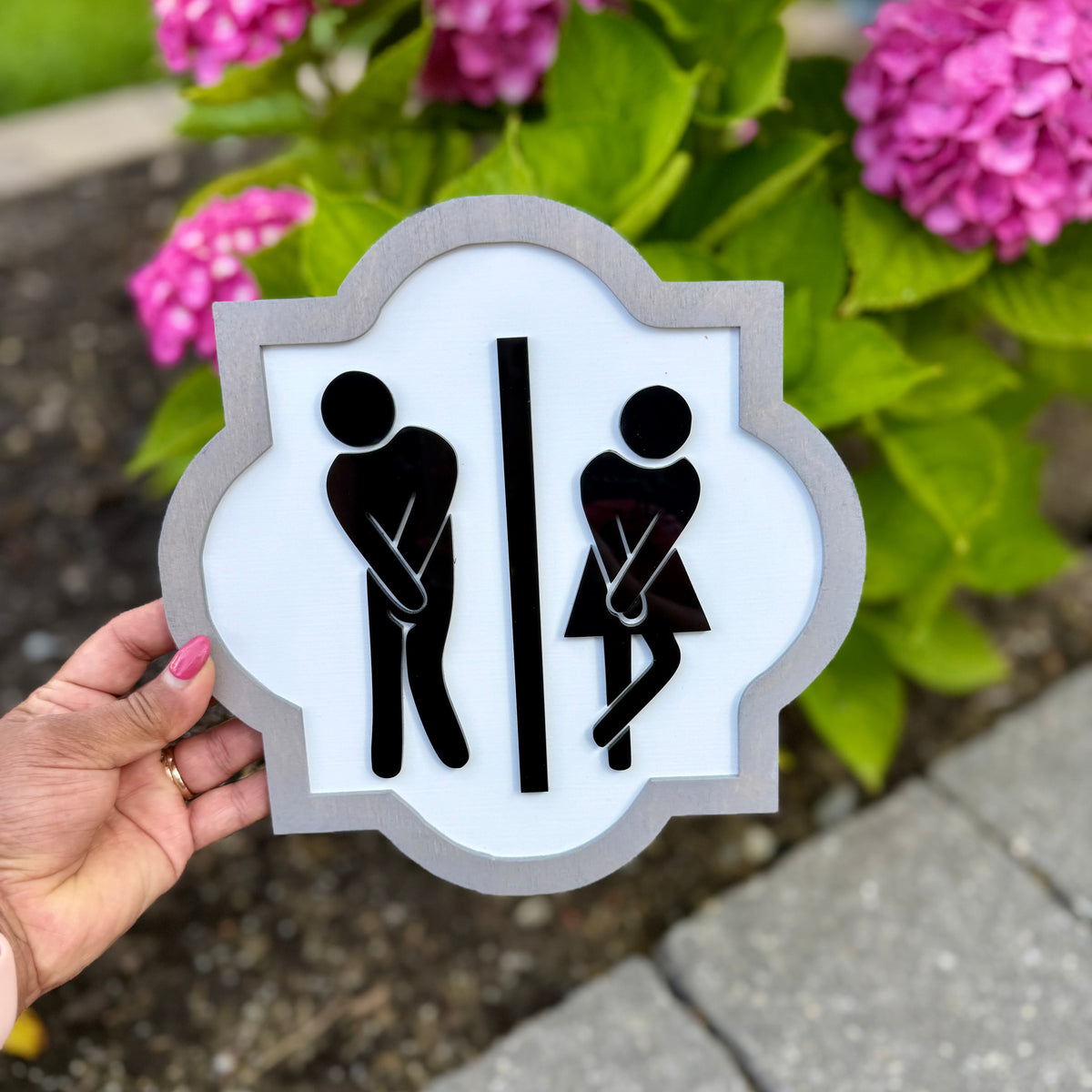 Hand holding a restroom sign with gender symbols in front of pink flowers.