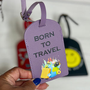 A hand with blue nail polish holds a lavender purple luggage tag with the words 'BORN TO TRAVEL' and a colorful suitcase graphic. The background is a white wooden surface with other blurred luggage tags.