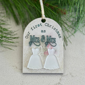 Decorative tag with 'Our first Christmas as Mrs & Mrs' text and wedding dress illustrations, hanging on a branch.