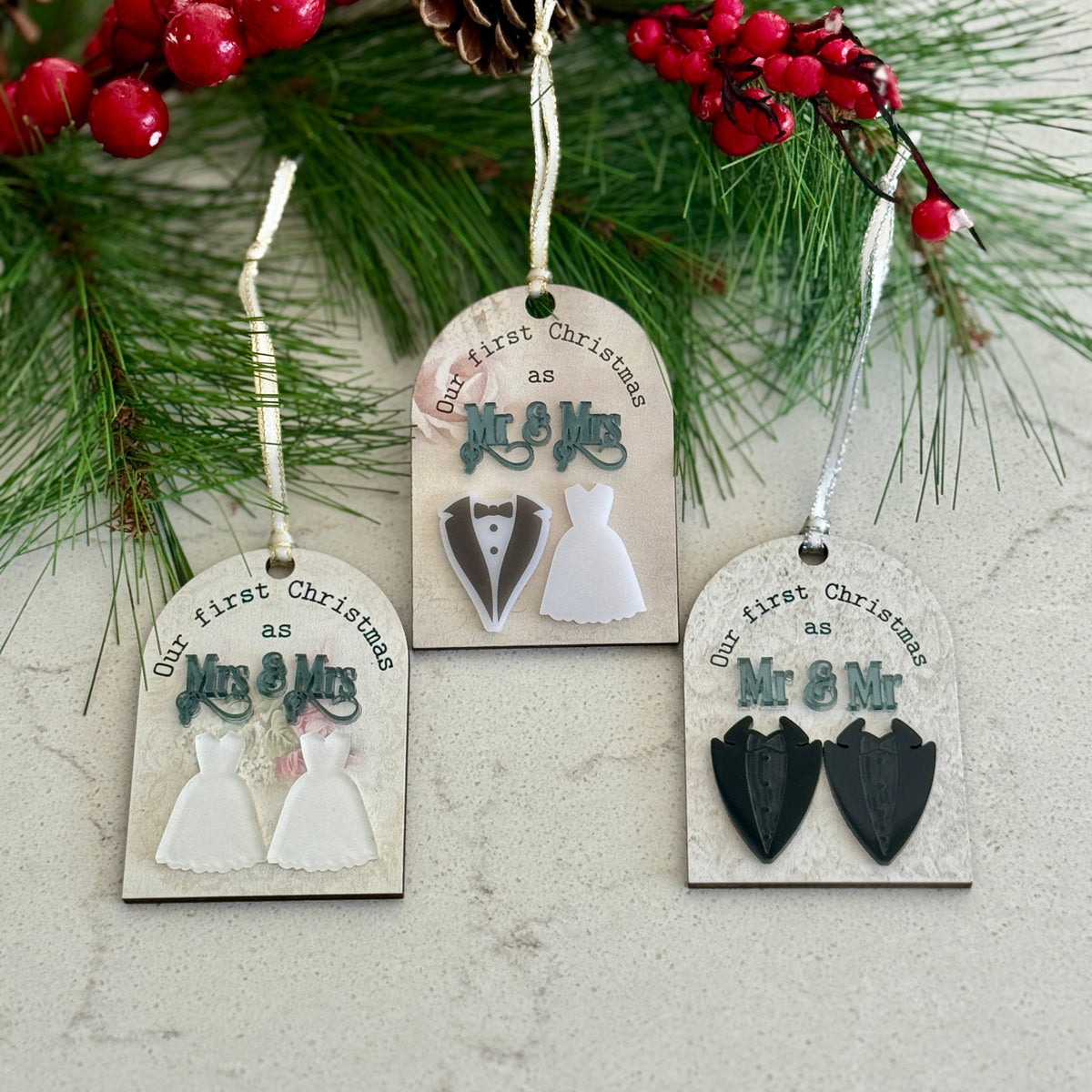 Three Christmas ornaments with wedding-themed designs on a branch with red berries and pine cones.