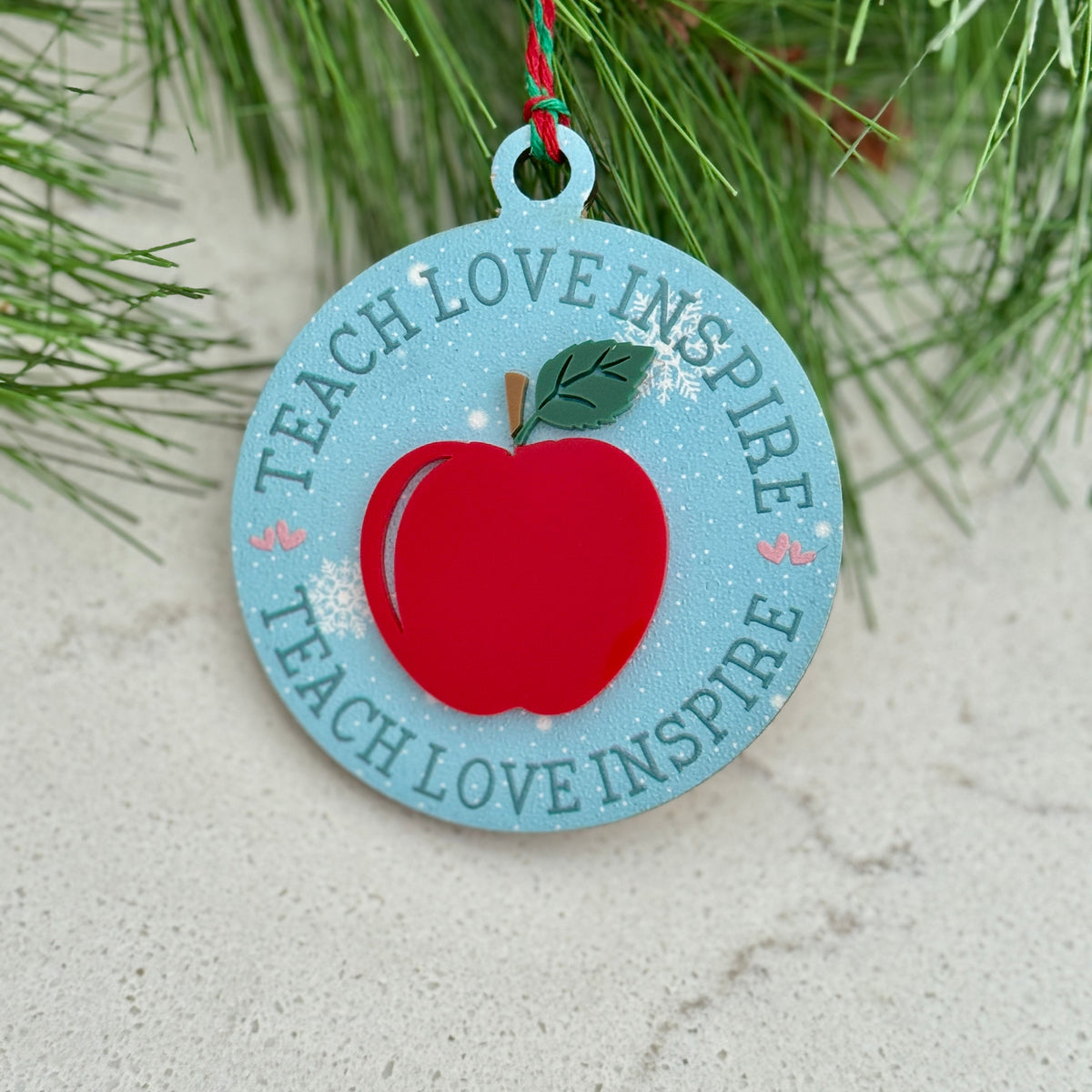 Decorative ornament with a red apple and 'Teach Love Inspire' text, hanging on a branch.