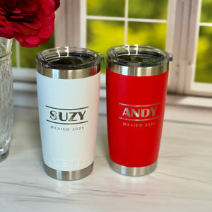 Two tumblers, one white and one red, with 'SUZY' and 'ANDY' branding on a surface with flowers in the background.