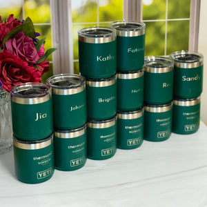 Set of green tumblers with names engraved, placed on a surface with flowers in the background.