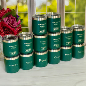 Stack of personalized green tumblers with names on a white surface with a blurred floral background