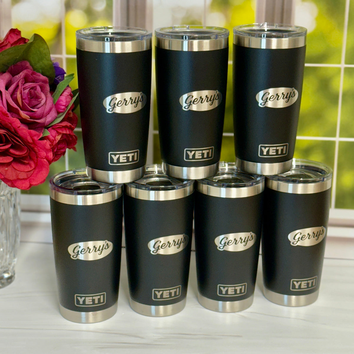 Set of black tumblers with silver lids and 'Gerry's' branding on a white surface with flowers in the background.