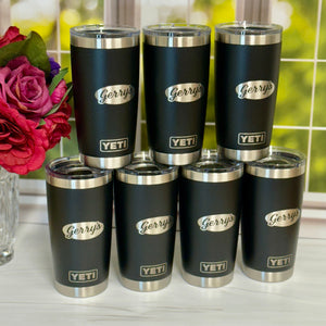 Set of black tumblers with silver lids and 'Gerry's' branding on a white surface with flowers in the background.