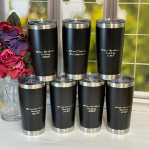 Set of black tumblers with silver lids on a surface with flowers and a window in the background.