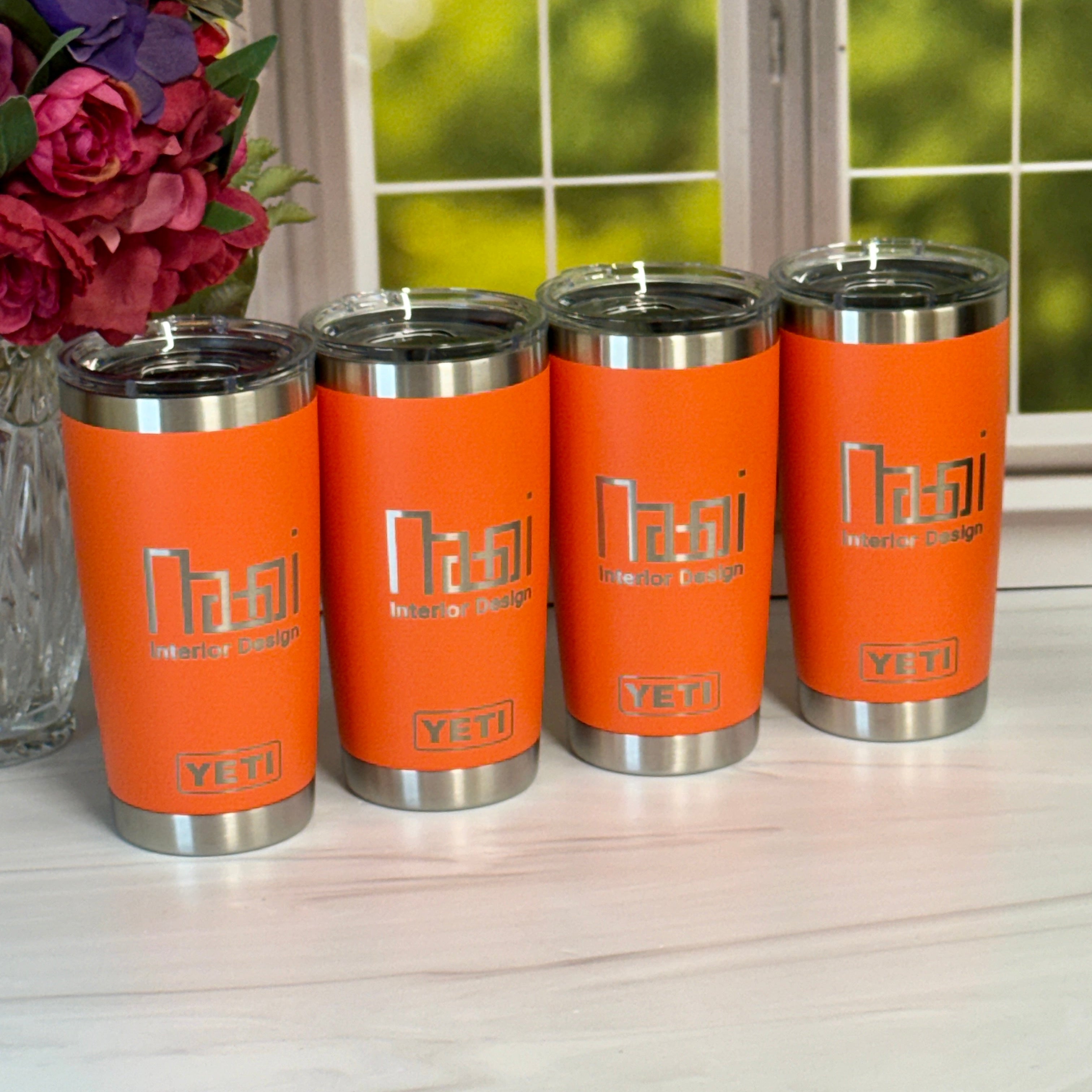 Four orange tumblers with silver lids on a light surface, with a blurred floral arrangement and window in the background.