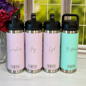Four YETI brand tumblers with personalized names on a surface with a blurred floral and window background.